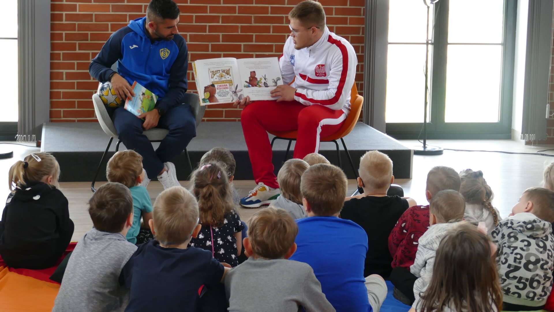 Na zdjęciu sportowcy czytający grupie dzieci siedzących w sali widowiskowej biblioteki