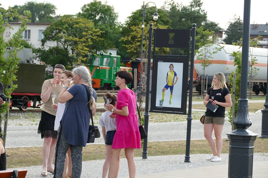 Na zdjęciu uczestnicy wystawy oglądający ekspozycję na tablicach na zewnątrz biblioteki. W tle lokomotywa i wagony