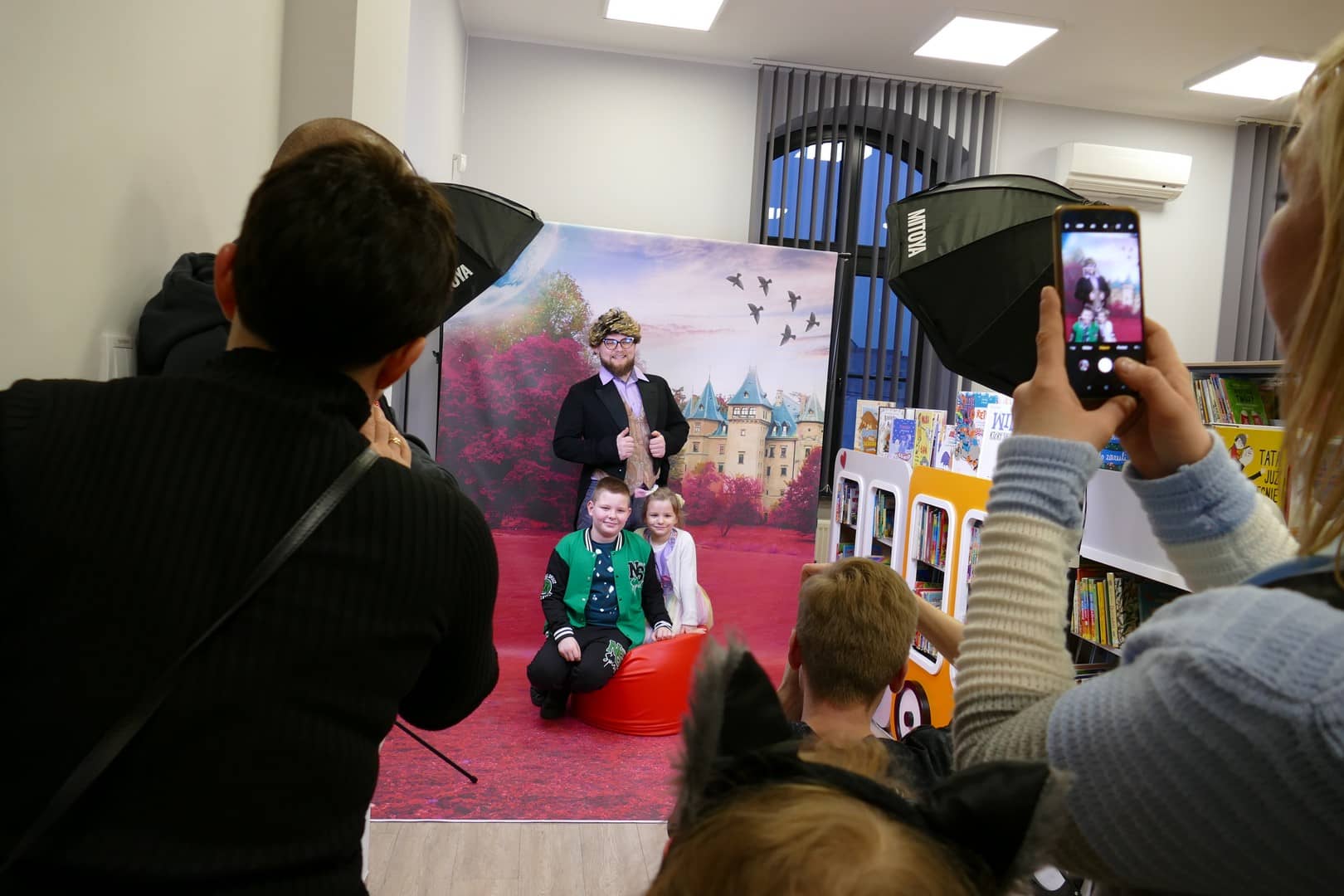 Na zdjęciu dzieci pozujące na ściance fotograficznej z animatorem przebranym za Pana Kleksa oraz rodzice wykonujący zdjęcia telefonami komórkowymi